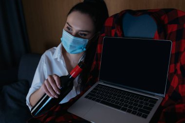Medikal maskeli üzgün kadın dizüstü bilgisayarın yanında boş ekranlı bir şişe şarap tutuyor.