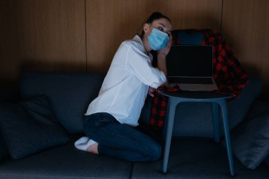 Medikal maskeli üzgün kadın dizüstü bilgisayarın yanındaki koltukta oturuyor. Sandalyesinde boş ekran var.
