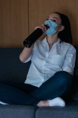 depressed woman in medical mask with hole drinking wine from bottle  clipart