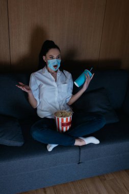 high angle view of shocked young woman in medical mask with hole watching tv with popcorn and soda at home clipart