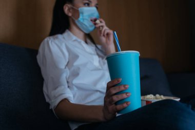 Elinde soda tutan ve evde televizyon izlerken patlamış mısır yiyen tıbbi maskeli genç bir kadının seçici odak noktası.