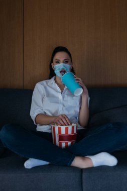 young woman in medical mask with hole sitting on sofa with crossed legs, drinking soda, eating popcorn and watching tv clipart