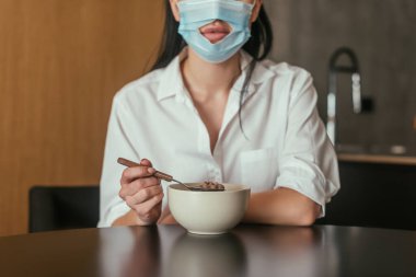 cropped view of woman in medical mask with hole for mouth holding spoon near bowl with breakfast clipart