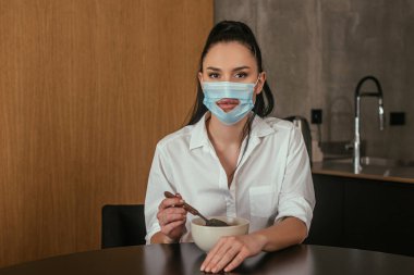 Delikli tıbbi maskeli genç bir kadın kahvaltı kaşığının yanında kaşık tutarken kameraya bakıyor.