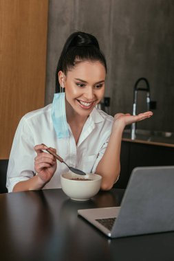 Kulağında koruyucu maskesi olan mutlu bir kız laptopta görüntülü sohbet sırasında kahvaltı yapıyor.