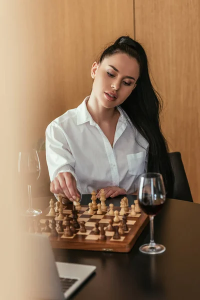 selective focus of attractive woman playing chess near laptop and red wine on table