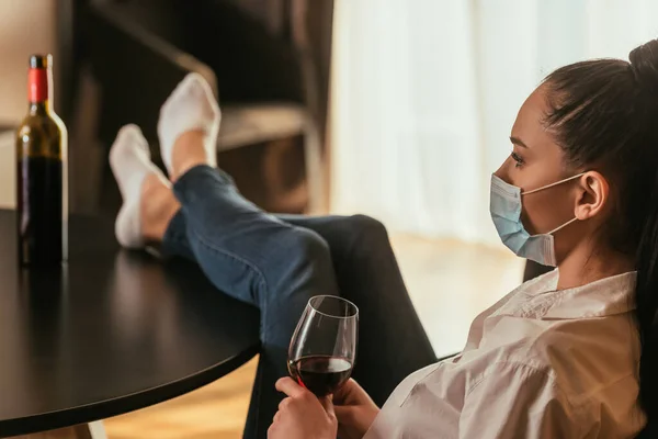 selective focus of depressed young woman in medical mask holding glass of red wine while sitting with legs on table