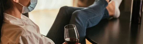 テーブルの上の脚で座っている間に赤ワインのグラスを持っている医療用マスクの女性のクロップドビュー 水平画像 — ストック写真