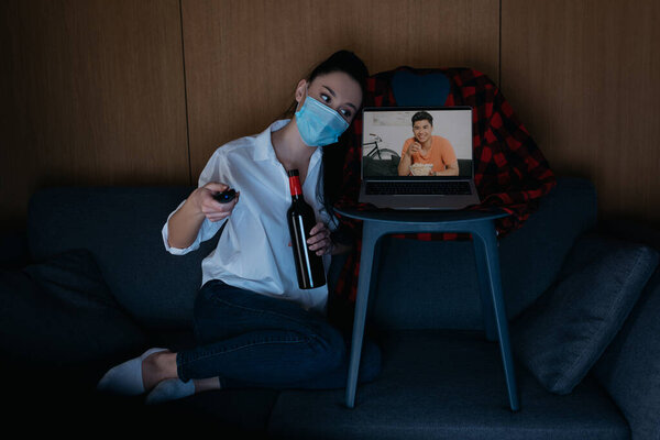 young woman in medical mask holding bottle of wine and tv remote controller while sitting on sofa near laptop with smiling asian boyfriend on screen