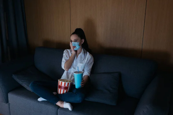 high angle view of worried young woman in medical mask with hole watching tv with soda and popcorn at home