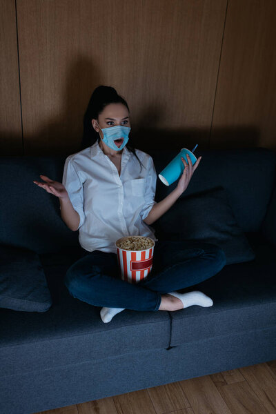 high angle view of shocked young woman in medical mask with hole watching tv with popcorn and soda at home
