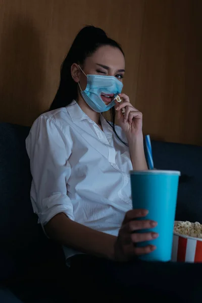 拿苏打水和吃爆米花时戴医疗面罩的年轻女性的选择性焦点 — 图库照片