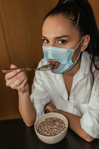 young woman in medical mask with hole for mouth having breakfast and looking at camera