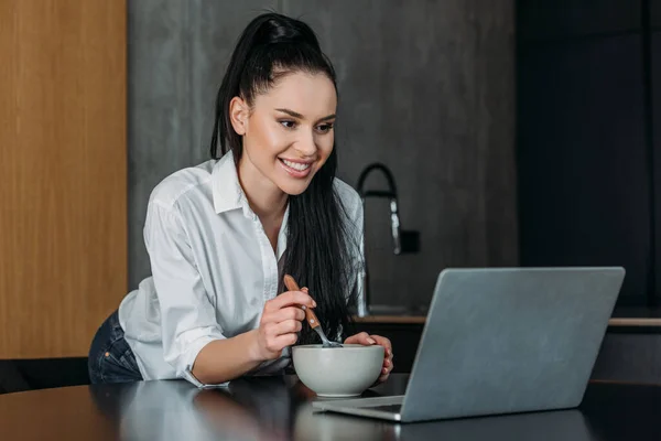 Mujer Feliz Mirando Computadora Portátil Mientras Sostiene Tazón Cuchara Cocina — Foto de Stock