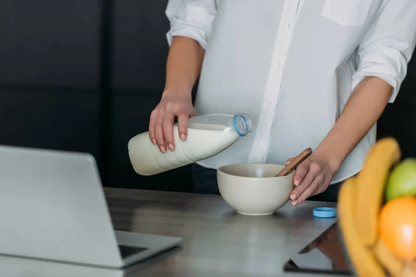 台所のノートパソコンの近くのボウルに牛乳を注ぐ女性のクロップドビュー — ストック写真