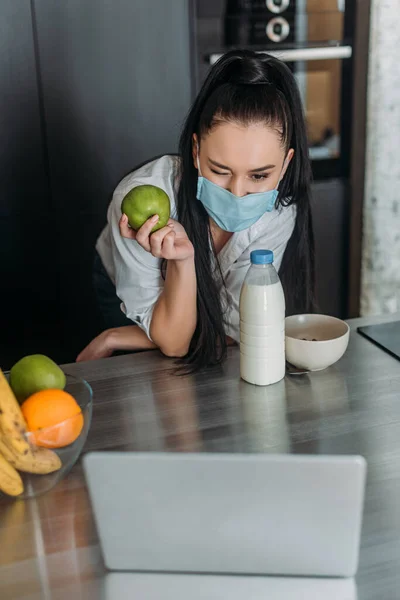 Mujer Joven Máscara Médica Sosteniendo Manzana Guiño Durante Videollamada Cocina —  Fotos de Stock