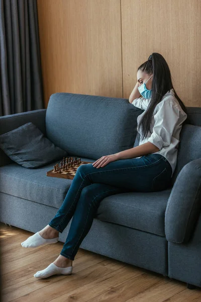 チェスボードの近くのソファに座っている医療用マスクの落ち込んでいる女性 — ストック写真