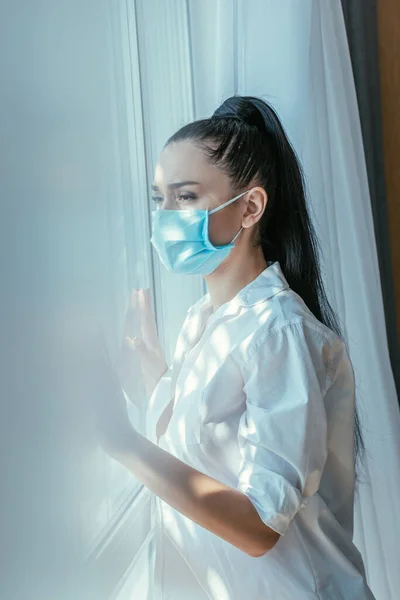 家庭で窓を通して見る医療マスクの悲しい女の子の選択的な焦点 — ストック写真