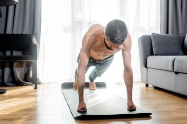 Oturma odasında spor paspası üzerinde antrenman yaparken tişörtsüz adam şınav çekiyor.