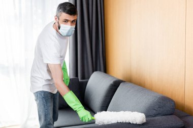 Tıbbi maskeli ve lastik eldivenli bir adam toz fırçasıyla kanepeyi temizliyor. 