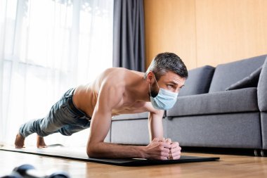 Medikal maskeli kaslı bir adamın evdeki spor minderinde kalas kullanmasının seçici odağı. 