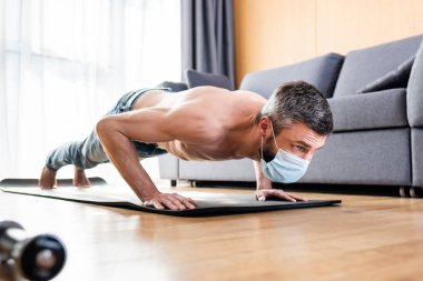Medikal maskeli üstsüz bir adamın oturma odasındaki spor minderinde şınav çekmesi.