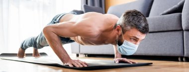 Sağlık maskeli üstsüz bir adamın panoramik mahsulü oturma odasındaki spor minderinde şınav çekiyor.