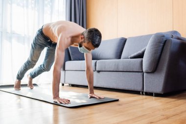 Sağlık maskeli gömleksiz adam evde spor yapıyor. 