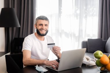 Elinde kredi kartı olan gülümseyen bir tele-işçi, tıbbi maske ve meyvelerin yanında dizüstü bilgisayar kullanıyor. 