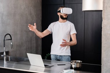 Bir adamın mutfak atölyesinde cihaz ve tıbbi maske yanında vr kulaklık kullanmasının seçici odağı 