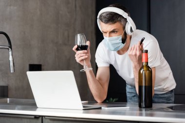 Kulaklıklı ve tıbbi maskeli dalgın bir adam USB bellek ve mutfakta dizüstü bilgisayarın yanında bir bardak şarap tutuyor. 