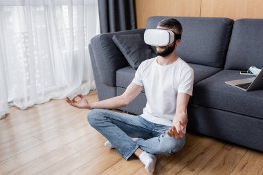 VR kulaklıklı adam oturma odasında lotus pozu vererek meditasyon yapıyor.