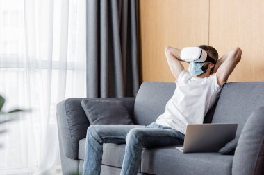 Tıbbi maskeli ve evdeki koltukta oturan sanal gerçeklik kulaklıklı adamın seçici odağı. 