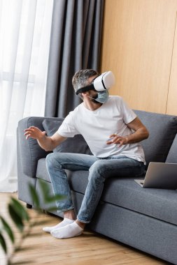 Tıbbi maskeli bir adamın oturma odasındaki koltukta VR kulaklığı kullanmasının seçici odağı.
