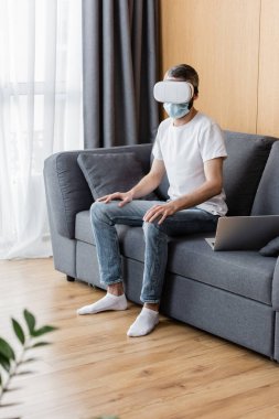 Tıbbi maskeli ve video kulaklıklı adamın seçici odağı kanepede dizüstü bilgisayarın yanında oturuyor. 