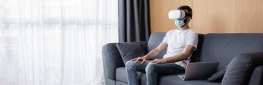 Tıbbi maskeli ve video kulaklıklı bir adamın kanepede dizüstü bilgisayarın yanında otururken panoramik görüntüsü. 