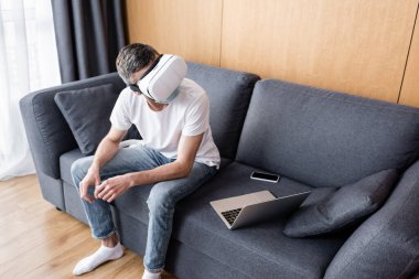 Video kulaklıklı ve tıbbi maskeli bir adam kanepenin üstünde akıllı telefon ve dizüstü bilgisayarın yanında oturuyor. 