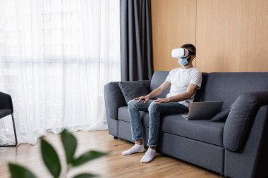 Oturma odasındaki kanepede dizüstü bilgisayarın yanında oturan VR kulaklıklı ve tıbbi maskeli adamın seçici odağı.