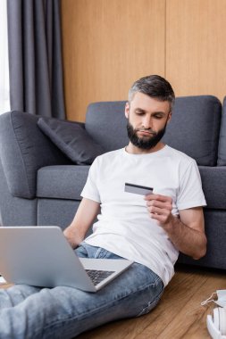 Sağlık maskesinin yanında kredi kartı ve dizüstü bilgisayar kullanan bir adam oturma odasında yerde kulaklık takıyor.