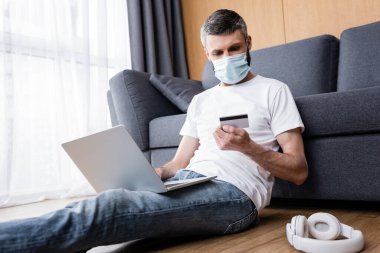 Maskeli bir adam oturma odasının zeminindeki kulaklıkların yanında dizüstü bilgisayar kullanırken kredi kartına bakıyor.