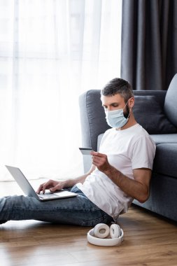 Maskeli bir adamın oturma odasındaki kulaklıkların yanında dizüstü bilgisayar ve kredi kartı kullanışının yan görüntüsü.