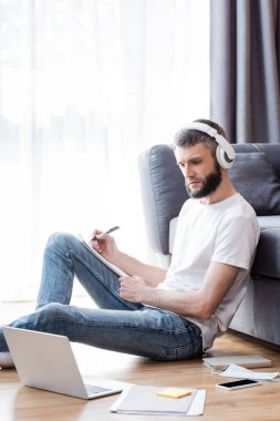Kulaklıklı adamın evde online eğitim sırasında dizüstü bilgisayar ve kırtasiye malzemelerinin yanına yazı yazışının yan görüntüsü