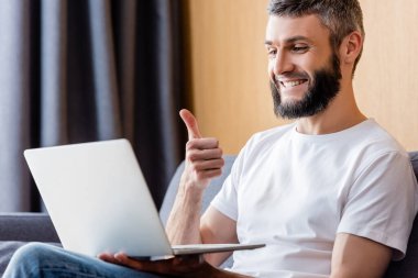 Evde dizüstü bilgisayarda video ararken baş parmağını gösteren neşeli serbest çalışanın seçici odak noktası 