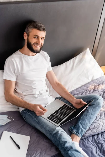 의료용 마스크와 침대에 노트북 근처에서 카드와 노트북을 미소짓는 사람의 — 스톡 사진