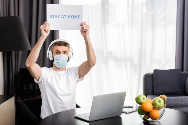 Freelancer in medical mask and headphones holding card with stay at home lettering near laptop and fruits on table