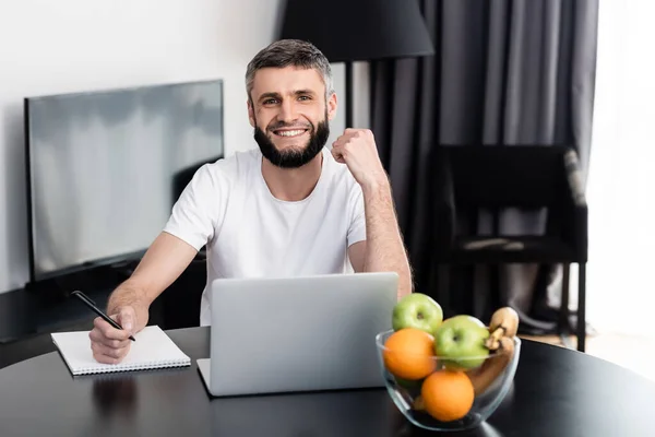 Handsome teleworker smiling at camera near notebook and laptop on table