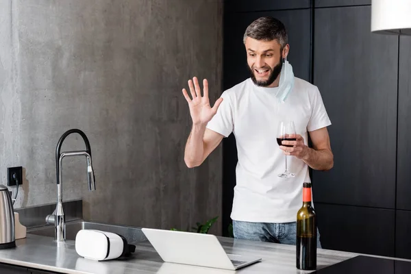 在厨房里一边拿着酒杯一边带着医疗面罩微笑的男人一边用笔记本电脑打电话 — 图库照片