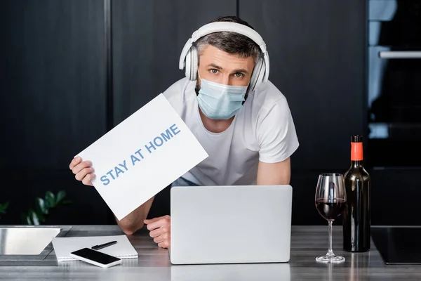 Man in medical mask and headphones holding card with stay at home lettering near wine and gadgets on worktop in kitchen