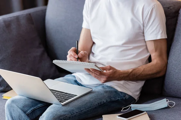 Vista Recortada Escritura Freelancer Cuaderno Cerca Máscara Médica Gadgets Sofá — Foto de Stock