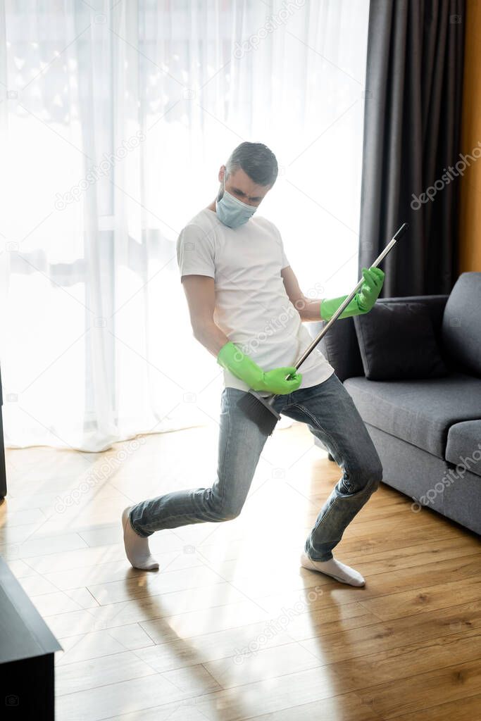 Man in medical mask and rubber gloves holding broom at home 